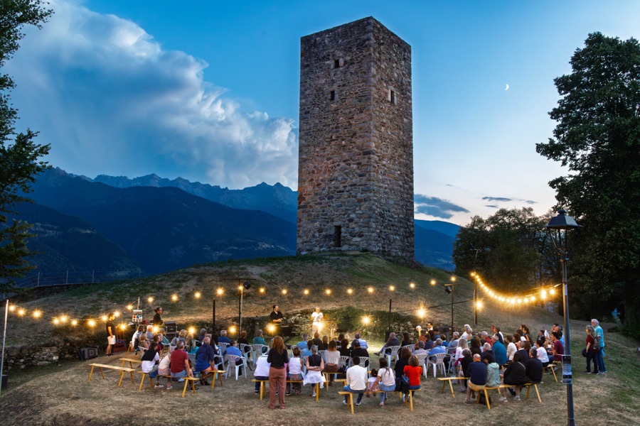Pubblico raccolto all'aperto ai piedi di una torre in pietra, al tramonto, tra le montagne, foto 17 di 17