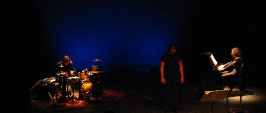 Tre figure in scena nel buio: percussioni, attore, pianoforte, foto 16 di 17