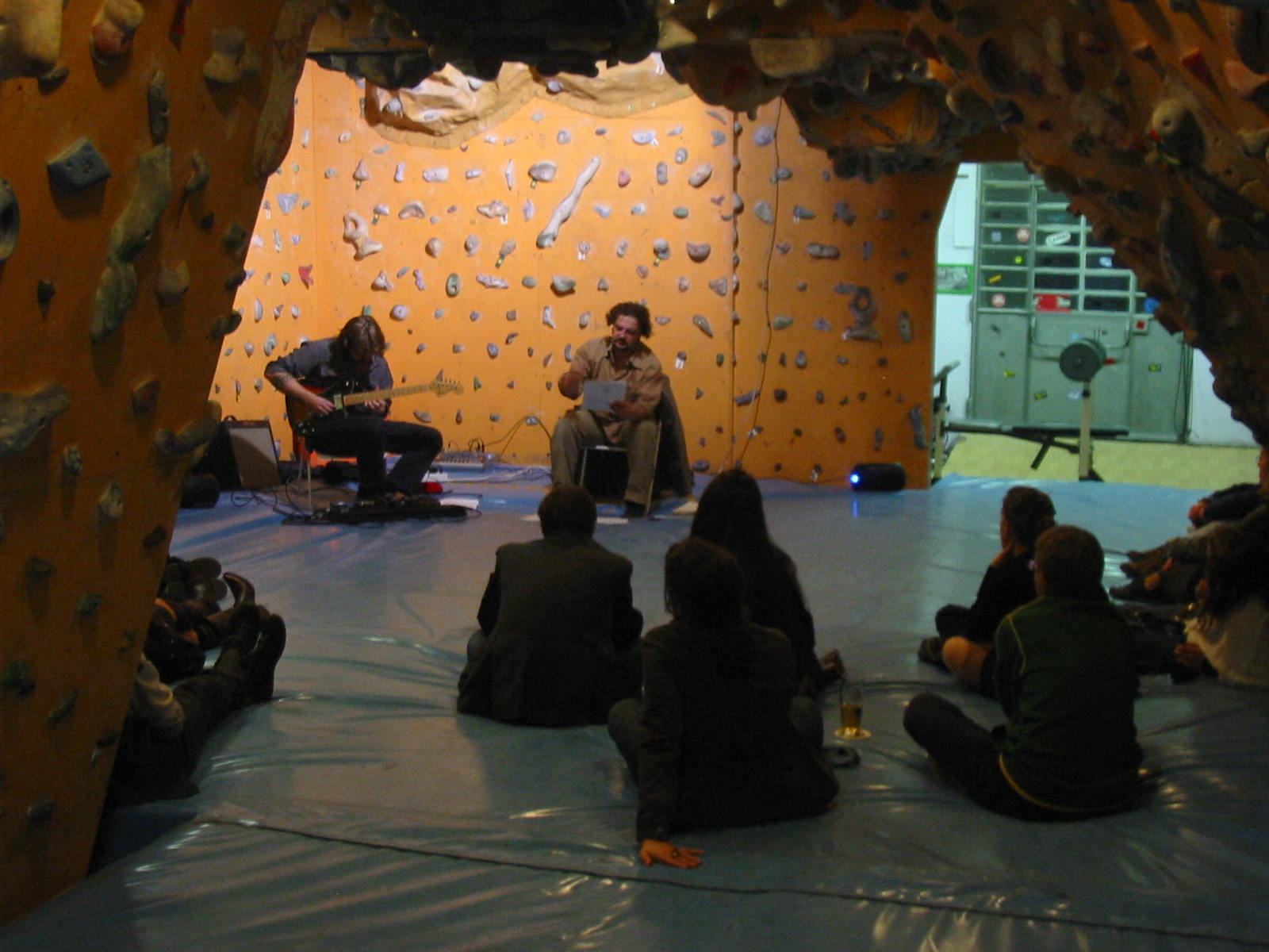 Un attore e un musicista seduti davanti a una parete da arrampicata, il pubblico a terra